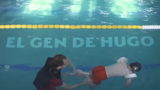 Frame del documental "El Gen de Hugo". Fotografía tomada en una piscina, en ella se ve a Hugo, un niño pequeño, nadando con la ayuda de su padre, que le sujeta las piernas para guiar los movimientos. Sobre ellos sobreimpresas las letras "EL GEN DE HUGO"